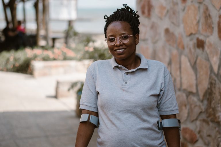 Nadia, a 33-year-old woman with a disability, participated in a learning event in Burundi. She is holding crutches and wearing a light grey t-shirt with black trousers and glasses.